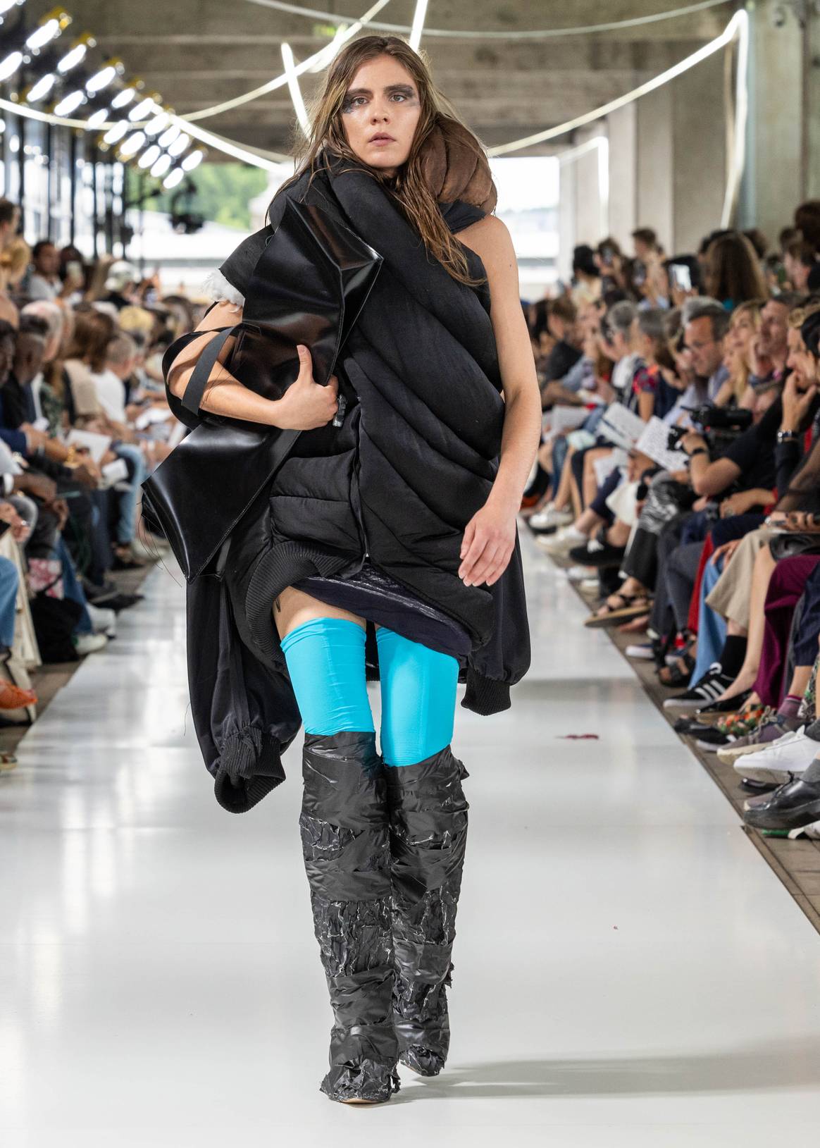 Woman on catwalk wearing padded black boots with bright blue spandex on top and sleeveless jacket.  IFM GRADUATE CLASS 2023.