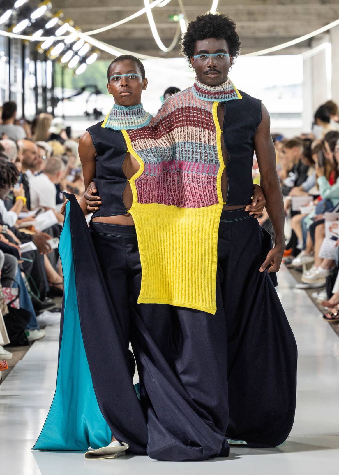 Man and woman stuck together through knitted fashion piece in yellow and red/blue/pink tones.